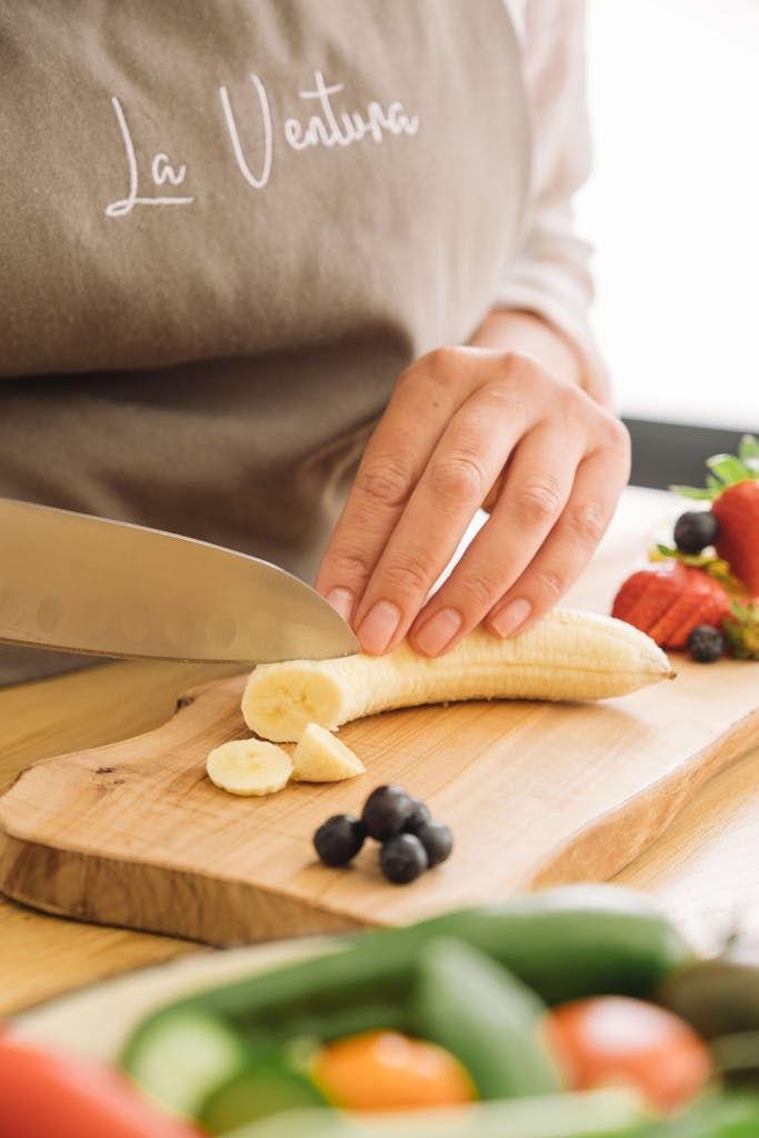A person slicing a banana on a wooden board with fresh fruits around.